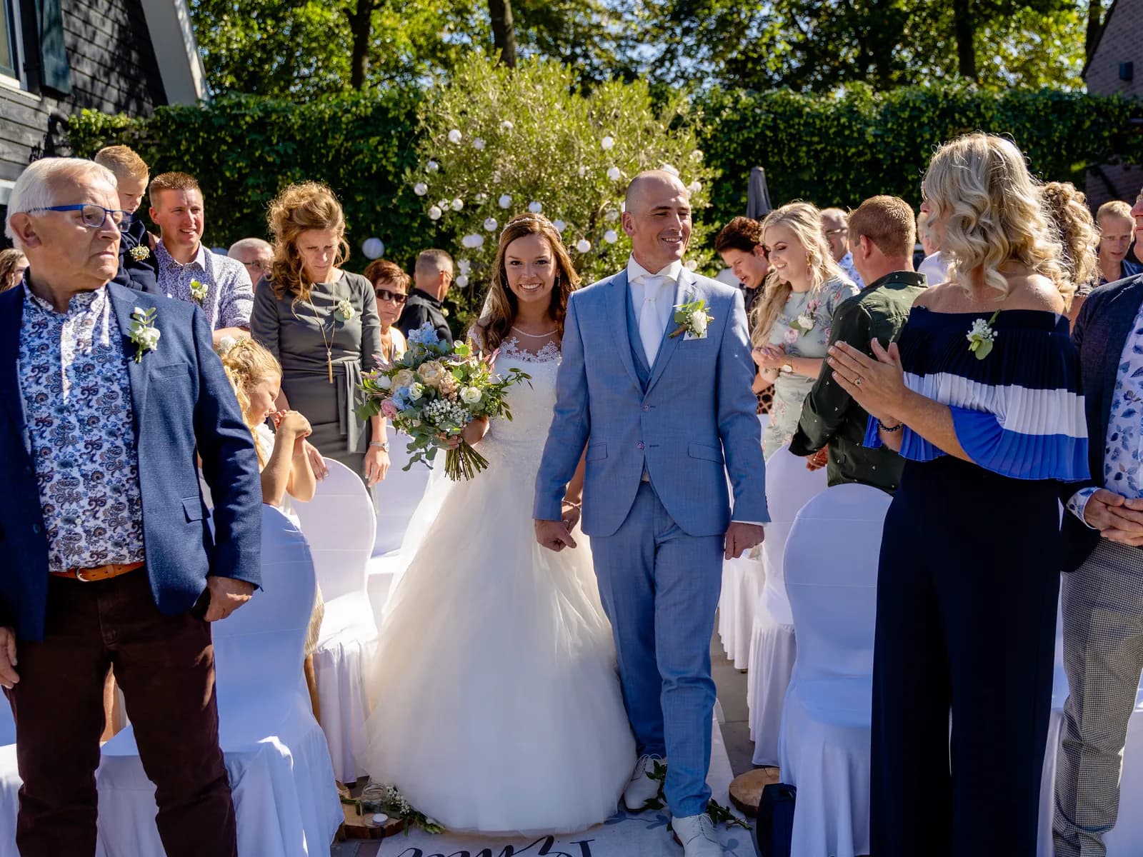 Bruidspaar loopt na de buitenceremonie hand in hand over het gangpad tussen de gasten