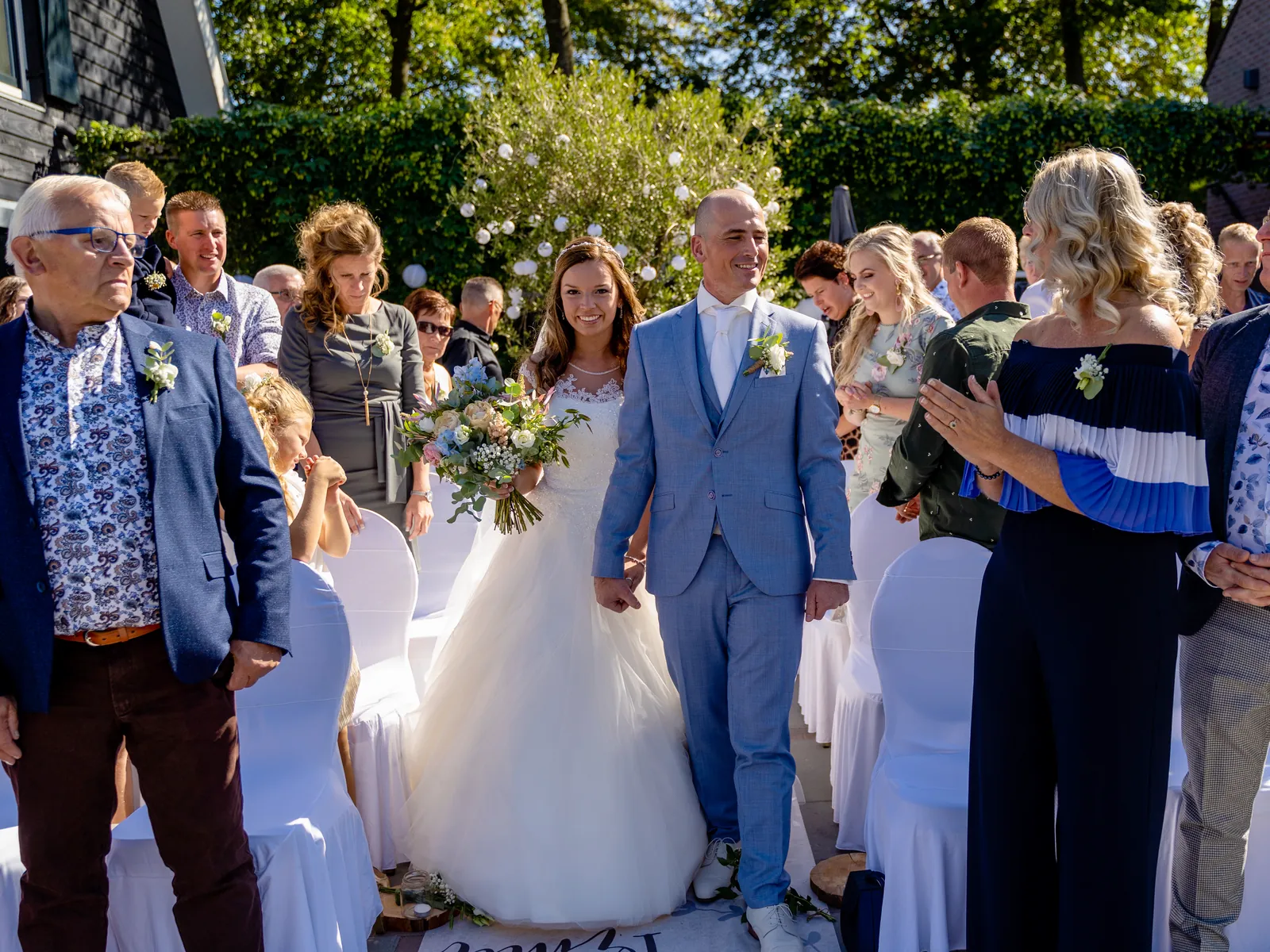 Bruidspaar loopt na de buitenceremonie hand in hand over het gangpad tussen de gasten